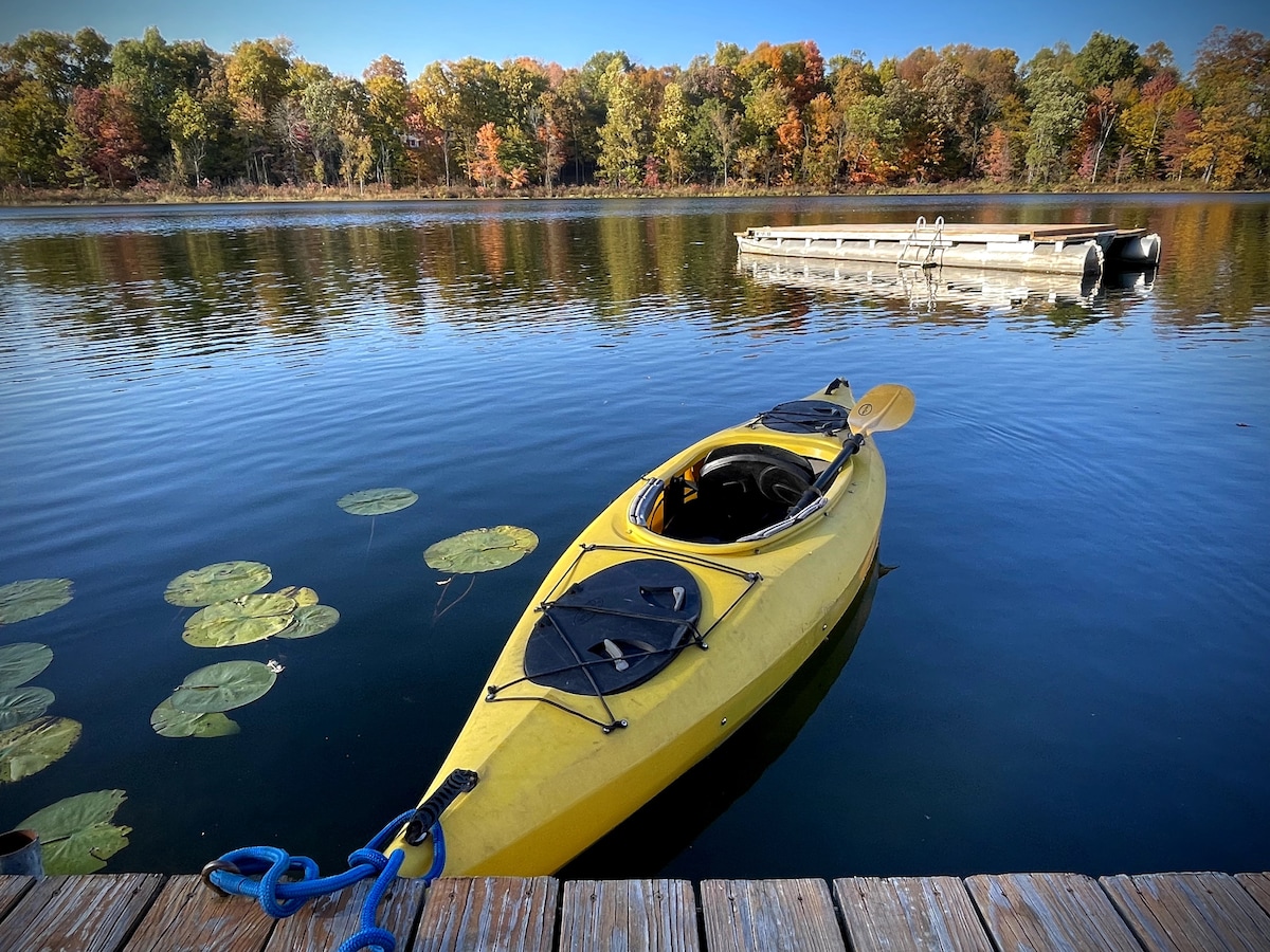 A bright yellow kayak is anchored near a wooden dock, floating gently on a serene lake. The water reflects a picturesque autumn landscape, with trees displaying vibrant fall colors in shades of orange and yellow. A swim platform is visible in the distance.