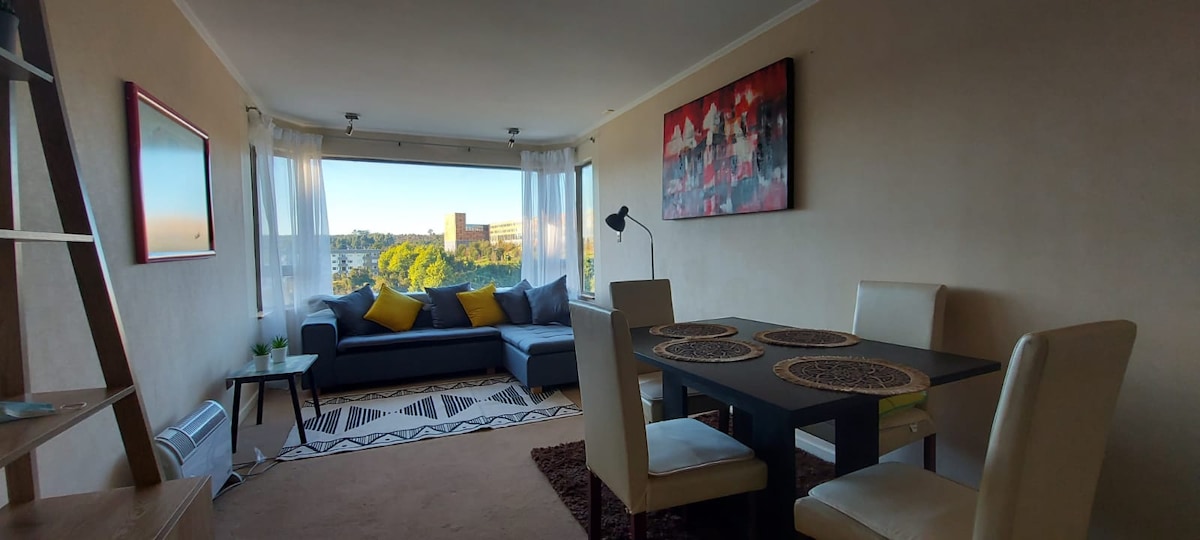 A bright living area features large windows offering a view of green surroundings. A gray sofa with yellow accent pillows complements a dark dining table surrounded by four light-colored chairs. An art piece adorns the wall, and a decorative rug adds warmth to the space.
