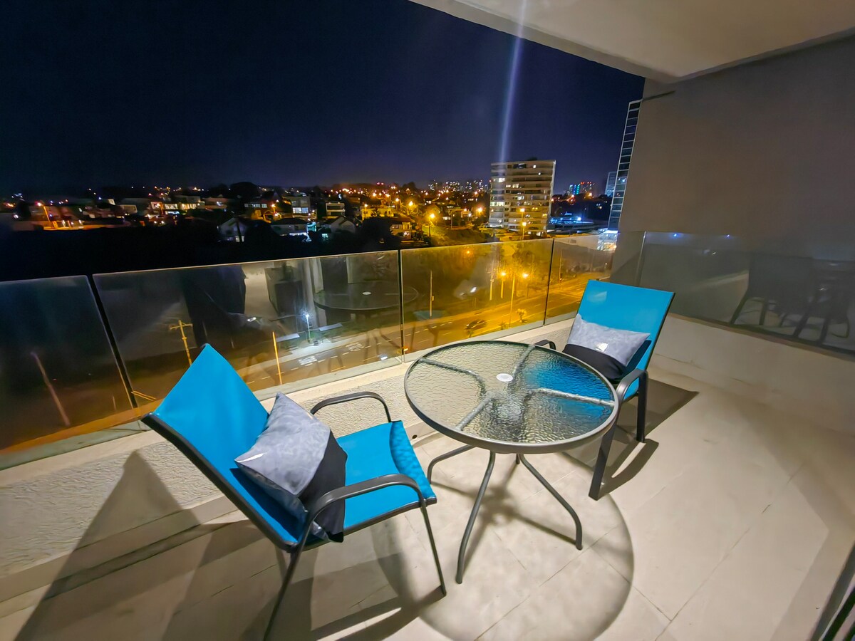 A balcony reveals a clear view of the city skyline at night, adorned by twinkling lights. Two blue chairs are positioned around a glass-topped table, providing a space for relaxation. A glass railing enhances the open feel of the balcony, inviting serene outdoor moments.