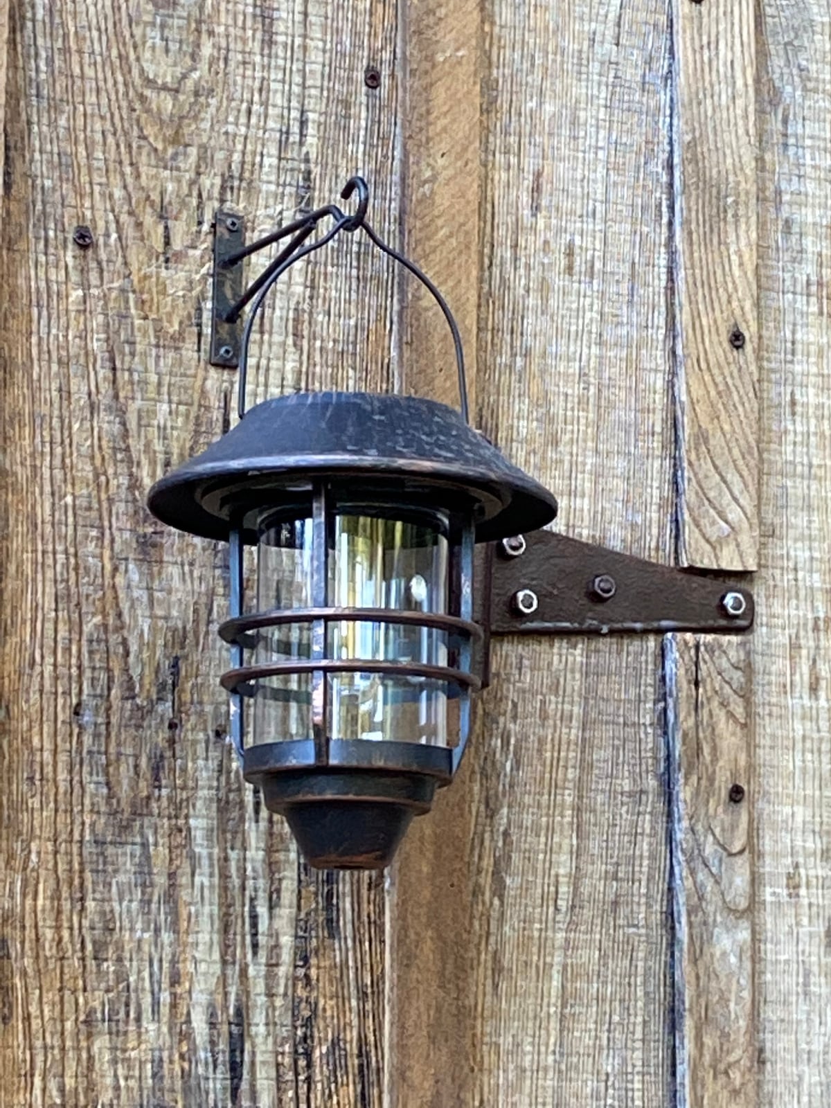 A rustic lantern is mounted on a wooden wall, featuring a metal frame with protective grating and a glass cover. The lantern is designed to provide illumination, adding a functional element to the cabin's exterior.