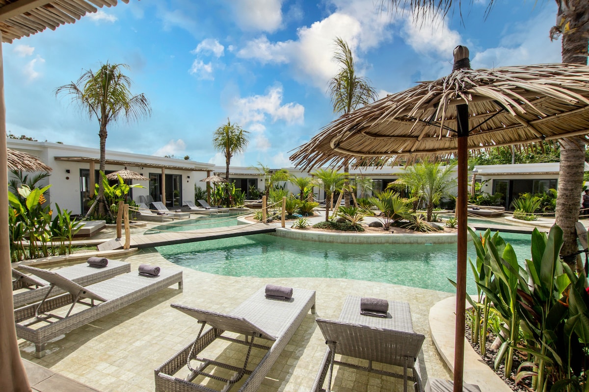 A lagoon-style pool is surrounded by lush tropical greenery and palm trees. Thatched umbrellas provide shade over comfortable lounge chairs placed near the water's edge. The scene features modern accommodations in the background, enhancing the tranquil outdoor space.