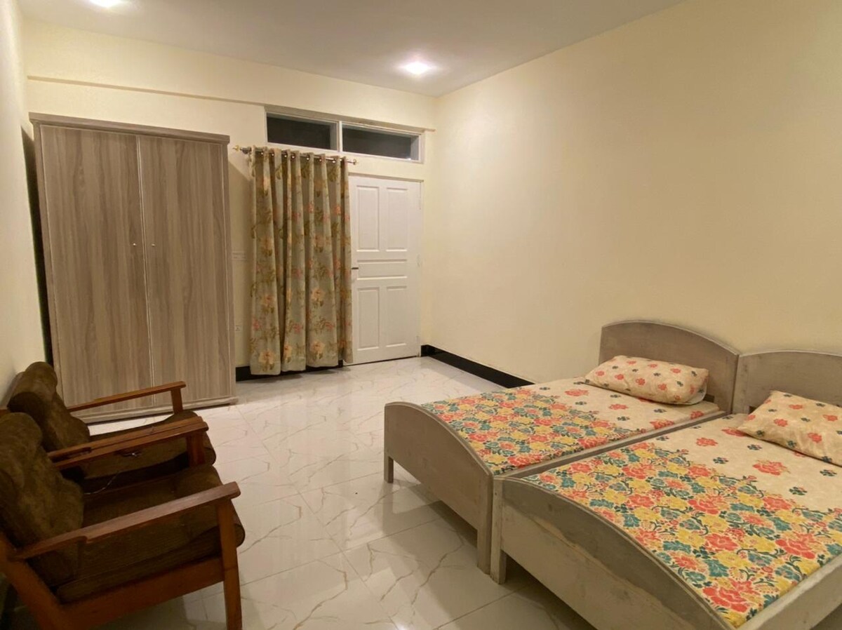 A spacious room features two beds, each adorned with floral-patterned bedding. A pair of wooden chairs offers additional seating. The room is illuminated by soft overhead lighting, and a wardrobe is positioned against one wall, complemented by window treatments that match the decor.
