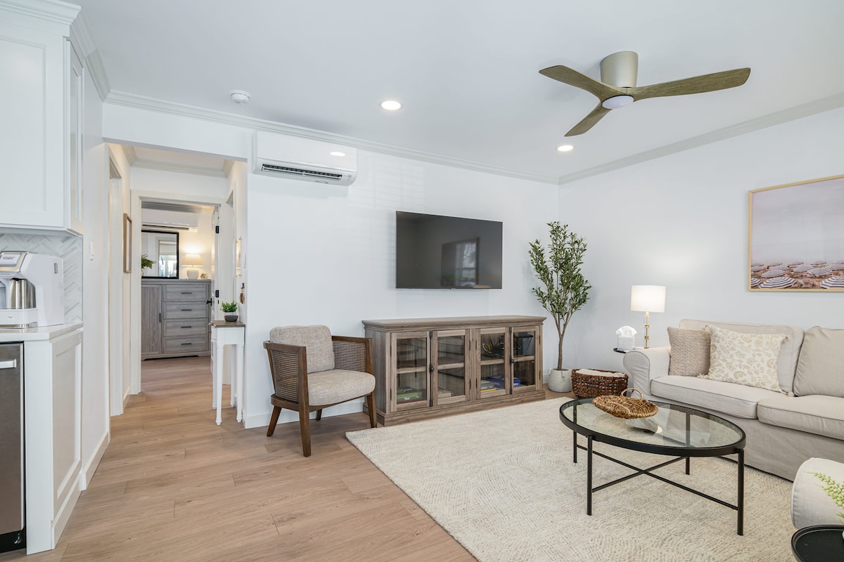 A cozy living area features a neutral color palette, complete with a light-colored sofa and a glass coffee table. A flat-screen TV is mounted on the wall, and a ceiling fan provides airflow. A plant adds a touch of greenery, enhancing the space's familiarity.
