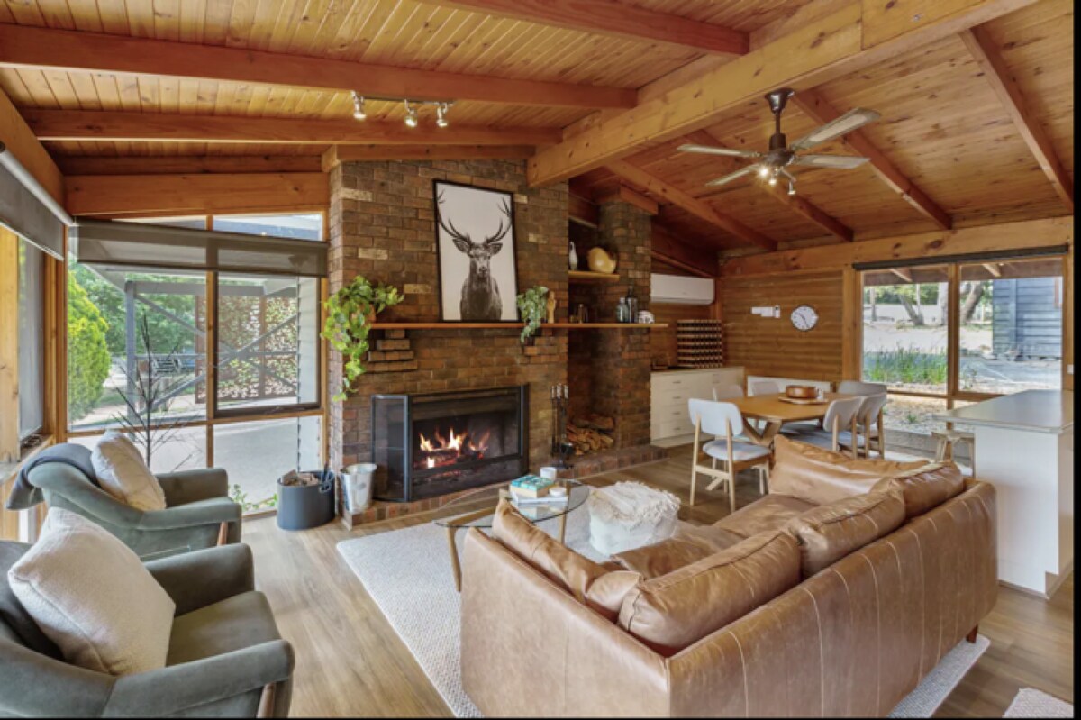 A spacious living area features wooden beams and a cozy fireplace, surrounded by comfortable seating. A large window allows natural light to fill the room, highlighting a carefully placed rug and tasteful decor, including a framed artwork above the fireplace.