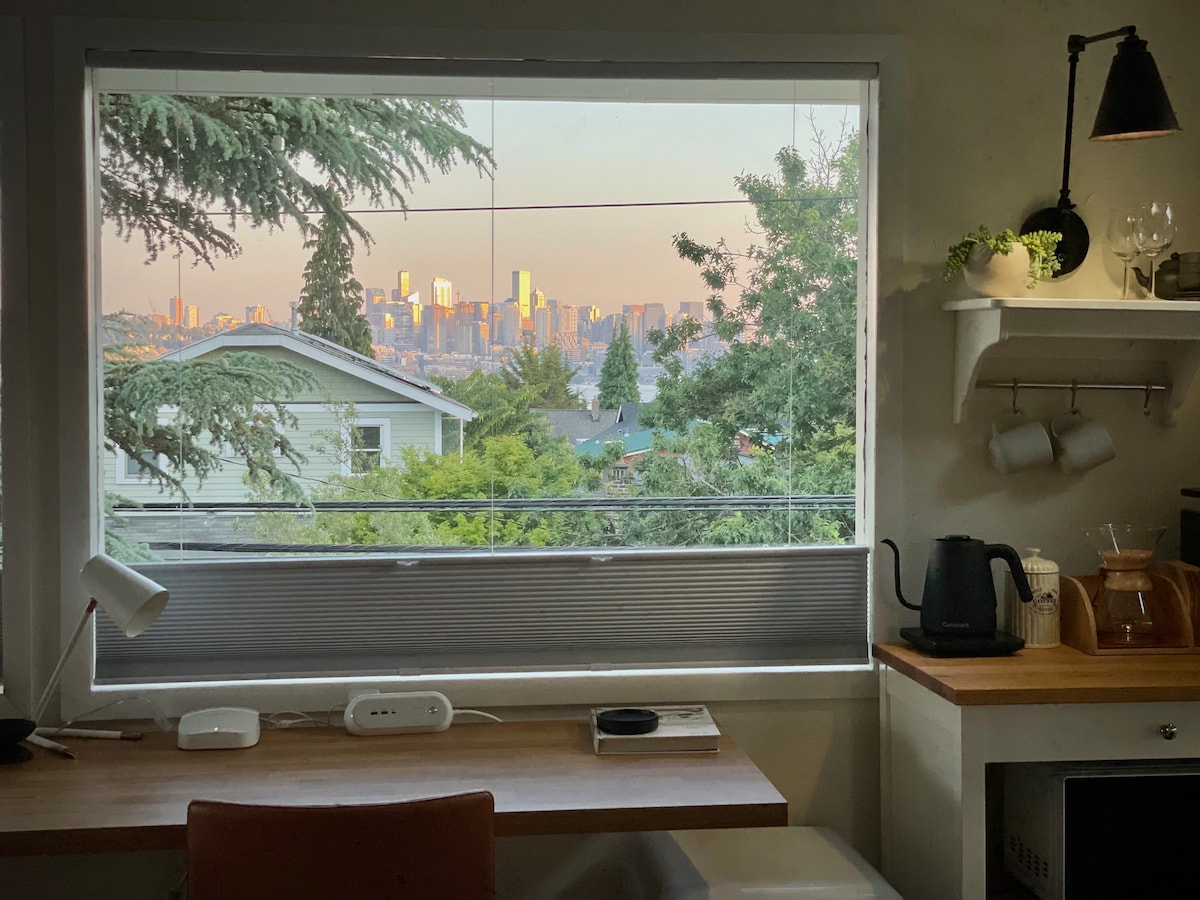 A cozy workspace is positioned beside a window, offering views of the Seattle skyline at sunset. Lush greenery is visible outside, while a kettle and wooden countertop complement the setup. Natural light floods the area, enhancing the tranquil atmosphere.