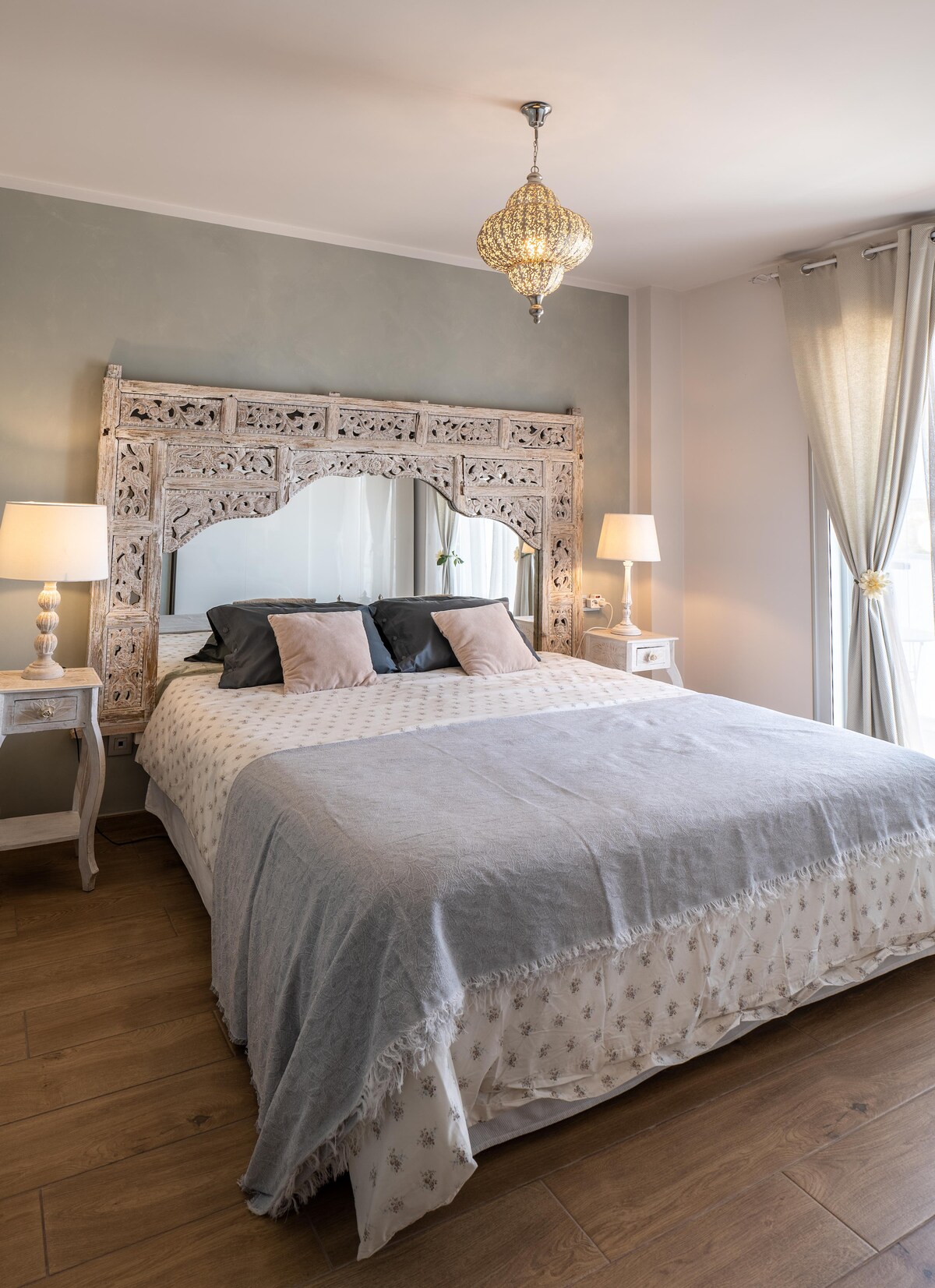 A double bedroom features a large bed with a decorative headboard and a light-colored blanket. Two bedside tables with lamps illuminate the space, complemented by elegant curtains and a softly shimmering chandelier above. Natural light enters through the window, enhancing the serene ambiance.