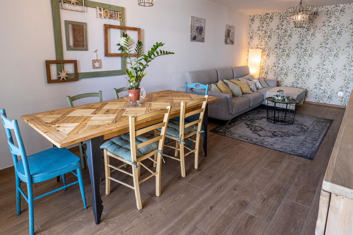 A dining area is furnished with a wooden table surrounded by colorful chairs. A cozy seating arrangement is visible in the background, featuring a grey sofa and decorative cushions. Natural light brightens the space, complemented by soft wall decor and a floral accent wall.