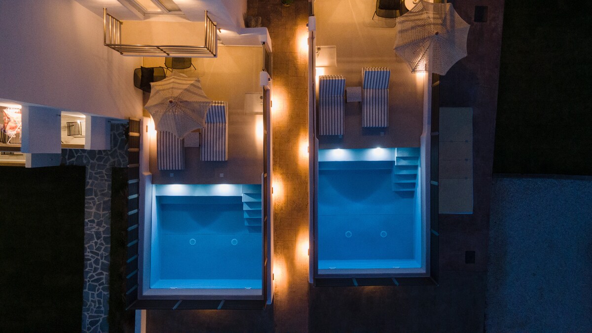 The image showcases two private pools illuminated in soft blue tones, surrounded by sunbeds and umbrellas. Warm outdoor lighting creates a serene atmosphere, highlighting the smooth textures of the pool edges and the surrounding patio area.