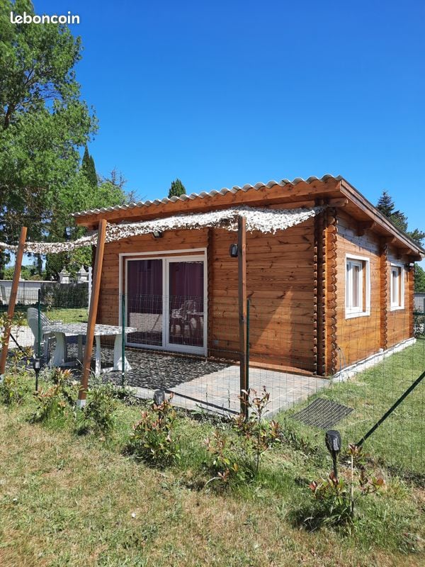 The exterior of a wooden chalet is displayed, featuring a sloped roof and terrace area. Large glass doors open to the entrance, framed by white window accents. Surrounding greenery adds to the inviting setting, with a landscaped yard and fence in view.