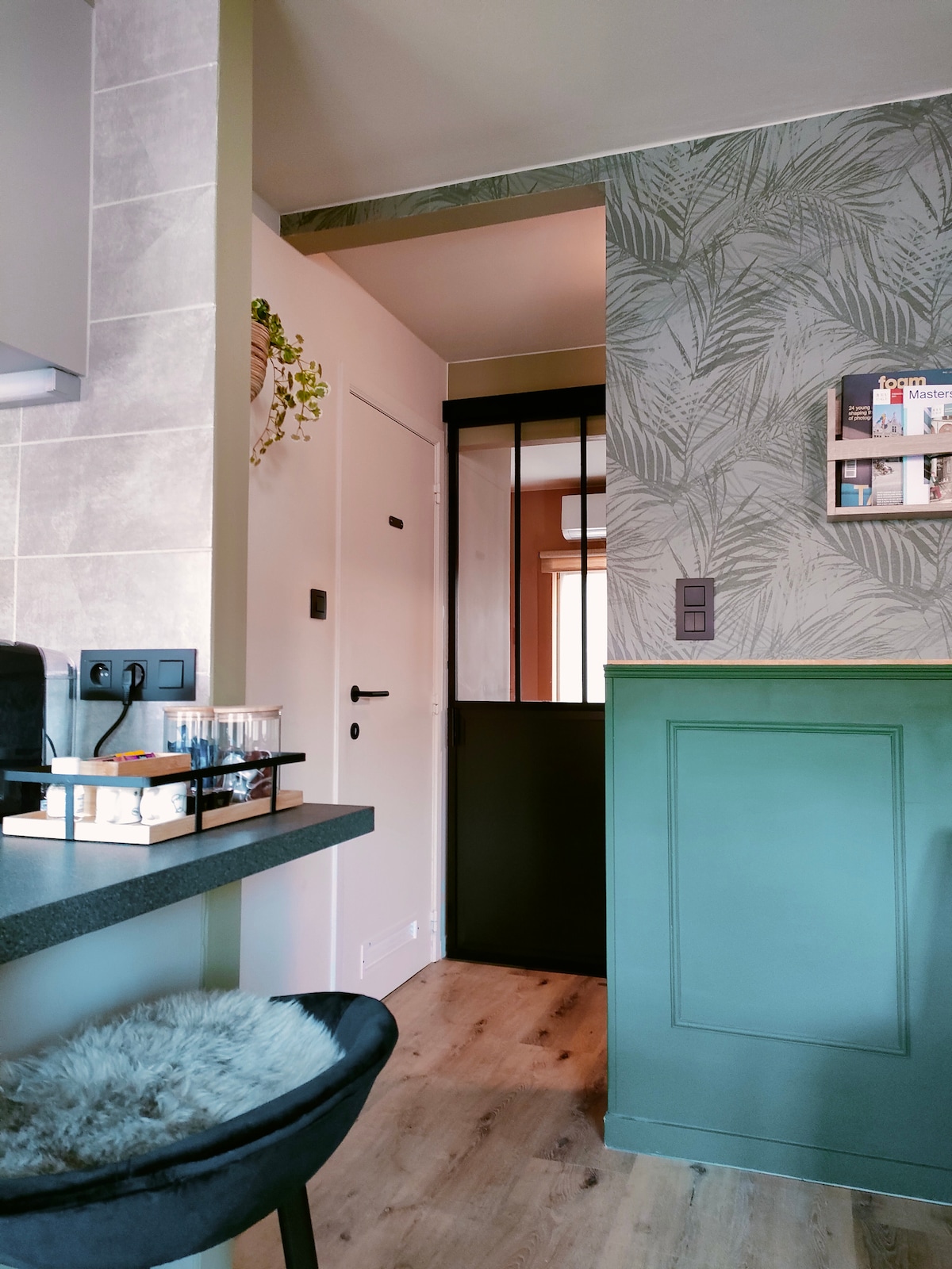 The open kitchen area features a sleek counter with a bar stool, complemented by greenery on the wall. A decorative sliding door leads to the entrance, with a touch of wallpaper displaying a botanical motif. Natural light enters the space, enhancing the inviting atmosphere.
