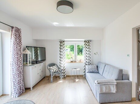 A bright living area features a gray sofa with a cozy throw blanket and a small round table. Natural light fills the space through a window, complemented by patterned curtains. A television is positioned on a white cabinet, while a wicker chair adds to the serene atmosphere.