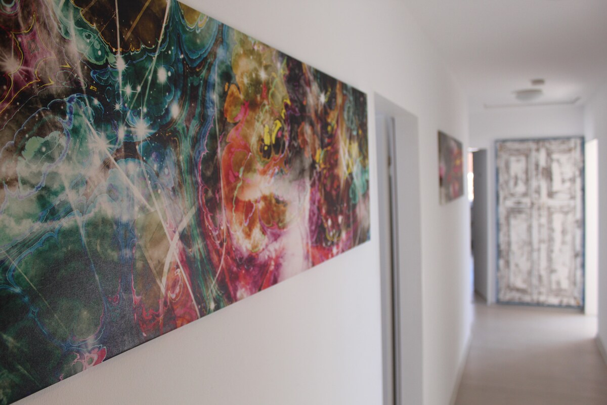 A bright hallway is lined with colorful abstract artwork, adding a splash of color to the white walls. The floor is tiled with a light, neutral color, and a door with a textured design leads to another room at the end of the corridor.
