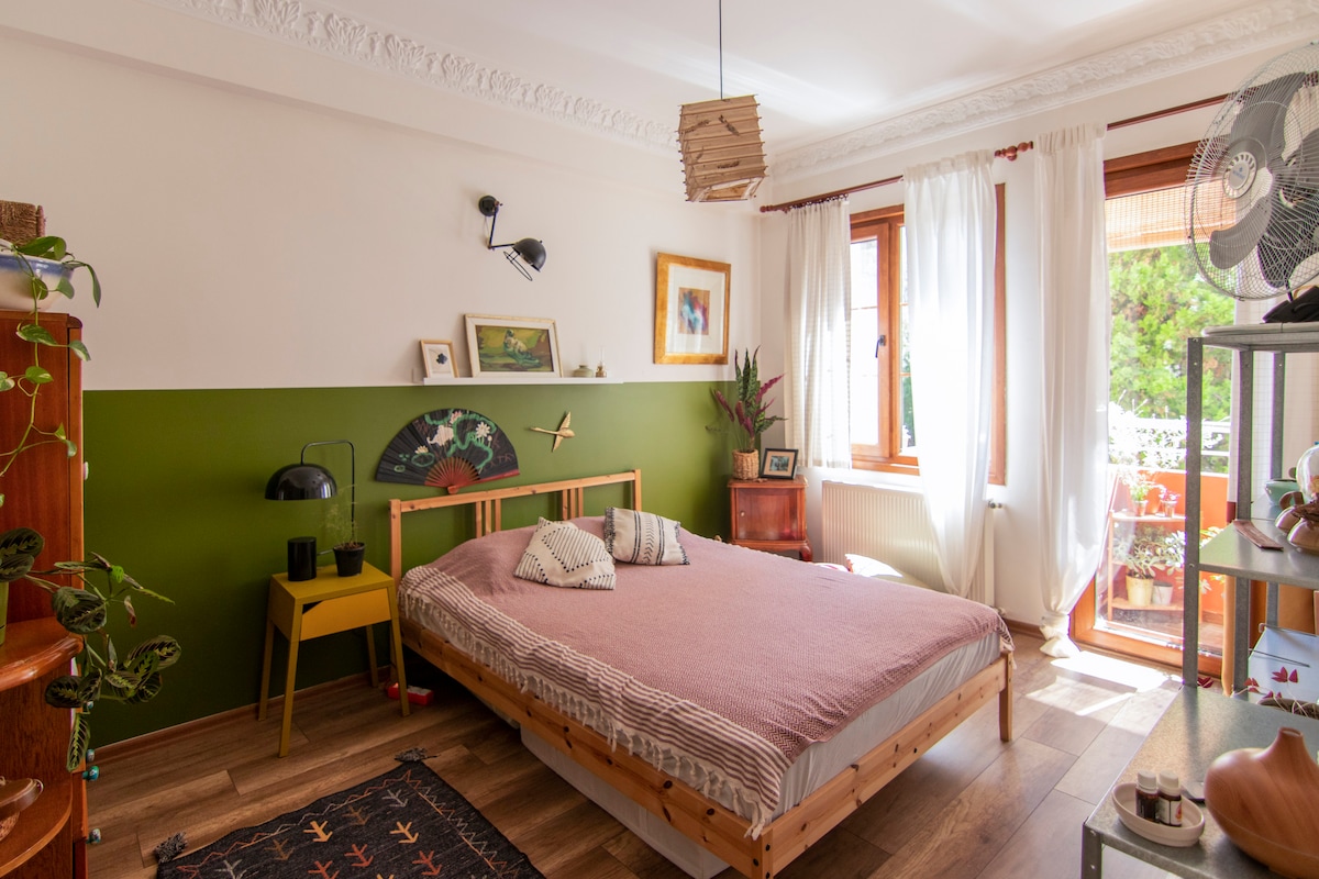 A spacious bedroom features a large wooden bed with decorative pillows and a green accent wall. Natural light flows in through large windows adorned with sheer curtains. Indoor plants and decorative pieces enhance the inviting atmosphere, completing the room's cozy design.