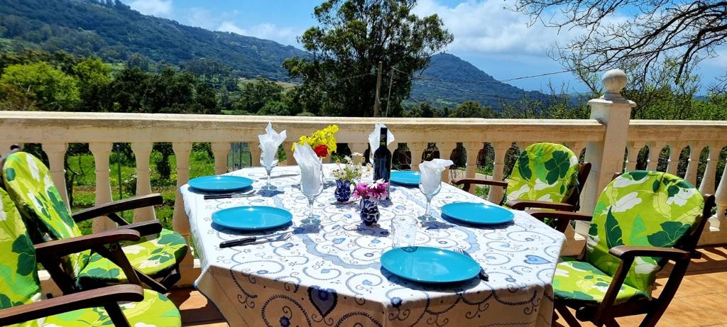 A dining table is set outdoors on a covered terrace, surrounded by lush greenery. Brightly colored chairs complement the blue plates and floral centerpiece. The mountains can be seen in the background under a partly cloudy sky, providing a serene dining atmosphere.