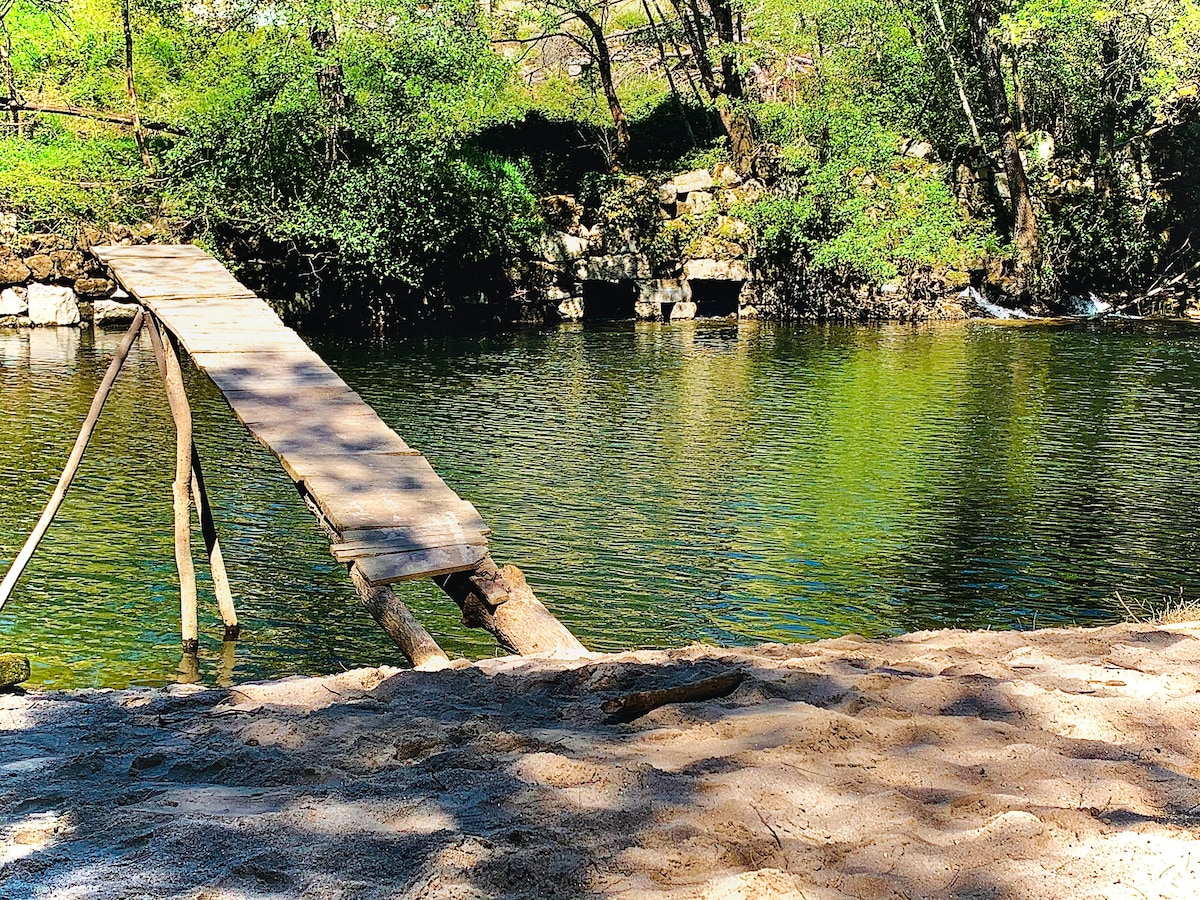 A rustic wooden bridge extends over a serene river, surrounded by lush greenery. The calm water reflects the vibrant colors of the landscape. Sand can be seen along the riverbank, providing a natural space for relaxation.