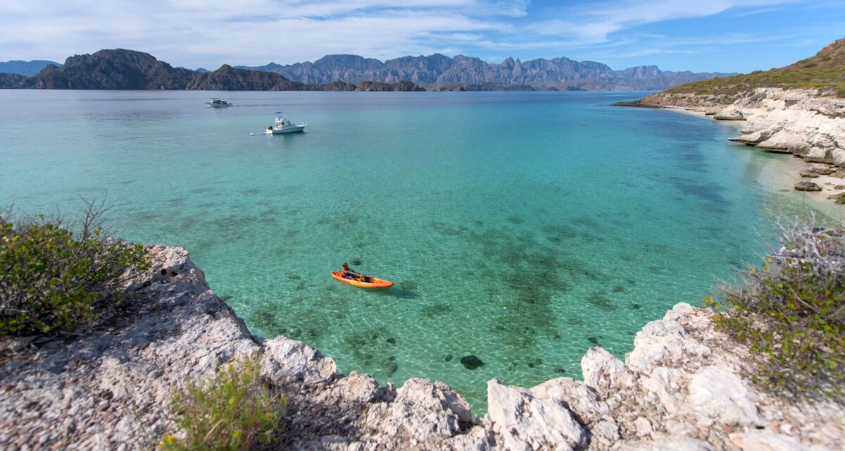 A serene coastal scene captures a kayaker in an orange kayak gliding through clear turquoise waters. The background features rocky cliffs and distant mountains, while a few boats can be seen anchored in the tranquil bay. Lush greenery lines the shore, contributing to the natural beauty.