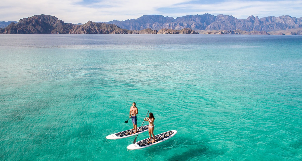 Two individuals are seen paddleboarding on clear turquoise waters, surrounded by mountainous landscapes in the distance. The calm ocean reflects the blue sky, creating a serene outdoor environment.
