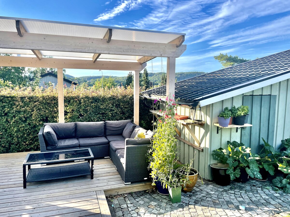 An outdoor seating area is showcased with a comfortable sectional sofa and a glass coffee table. Lush greenery surrounds the space, while a wooden pergola provides overhead shelter. Potted plants are arranged along the wall, contributing to the serene setting.