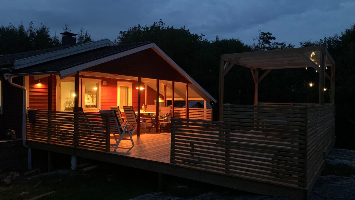 A cozy wooden cabin is illuminated at night, showcasing a spacious deck with comfortable seating. The area features a covered gazebo and outdoor lights, creating a welcoming atmosphere. Large windows provide a glimpse into the warm interior, emphasizing a connection to the outdoors.