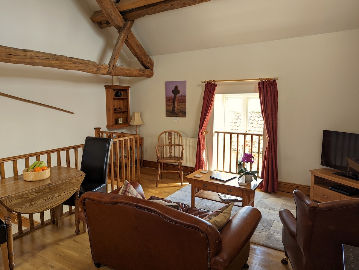 An open-plan living area features original oak cruck beams and high ceilings. Comfortable leather seating is arranged around a coffee table, with a smart TV positioned nearby. A round wooden dining table is paired with wooden and upholstered chairs. Natural light floods in from a window, adorned with curtains.