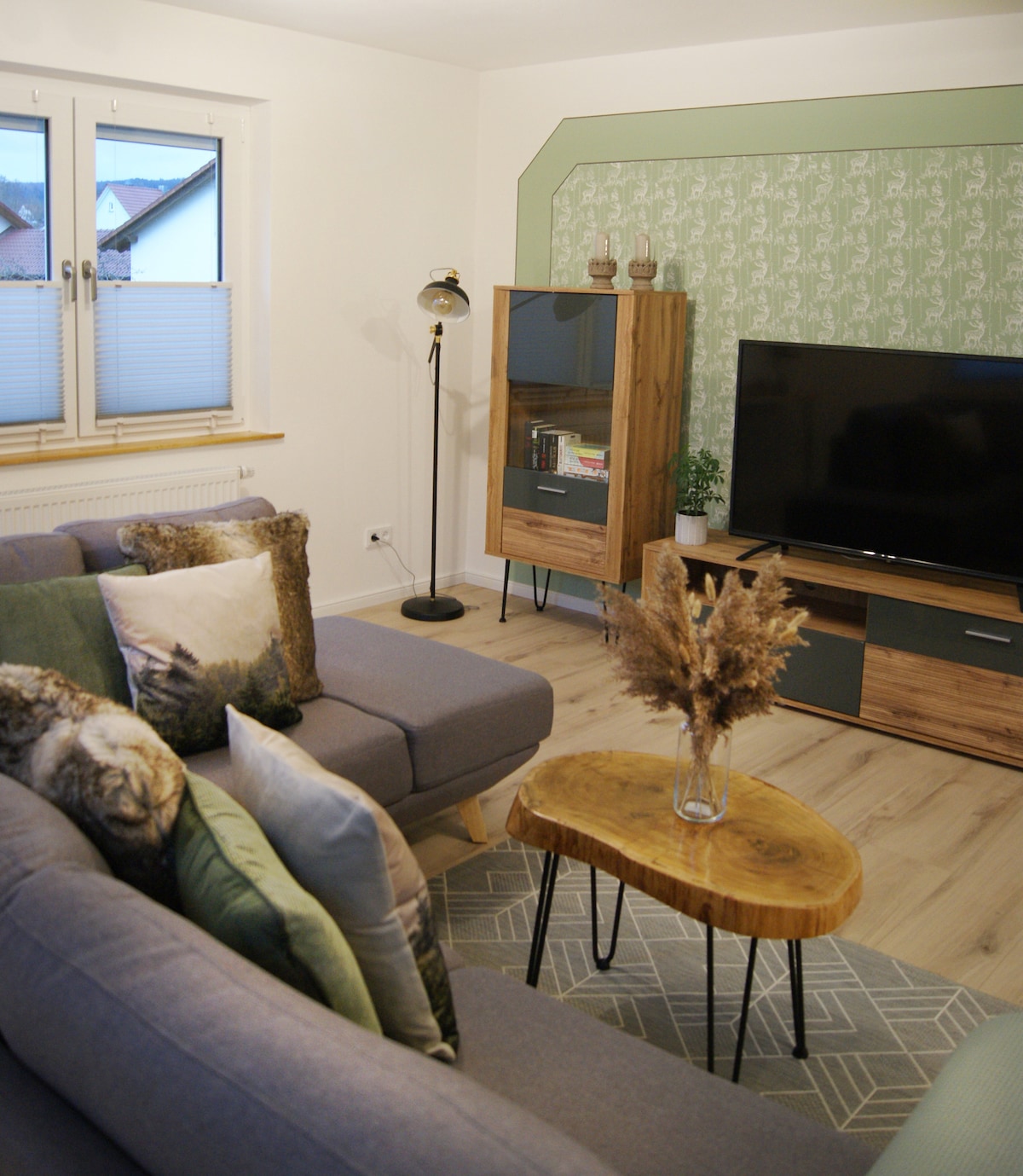 A modern living area features a pair of comfortable sofas adorned with decorative cushions. A wooden coffee table is centered between the sofas, complementing the light wood floors. A TV cabinet and an accent cabinet are positioned against the wall, with natural light streaming in from two windows.