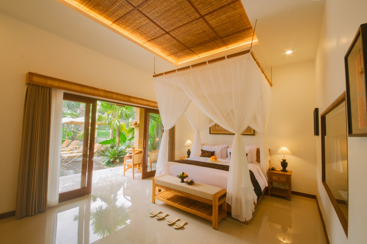 A serene bedroom is highlighted by a four-poster bed draped with sheer fabric. Natural light floods the space through glass doors that open to a tranquil outdoor area with tropical greenery. Minimalist furnishings complement the calming ambiance.