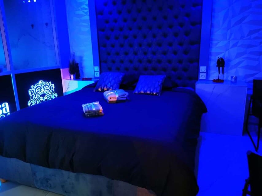 A spacious bed with a tufted headboard is positioned against a geometric wall. Soft blue lighting envelops the room, creating a calming ambiance. Two decorative pillows rest on the bed, accompanied by folded blankets at the foot.