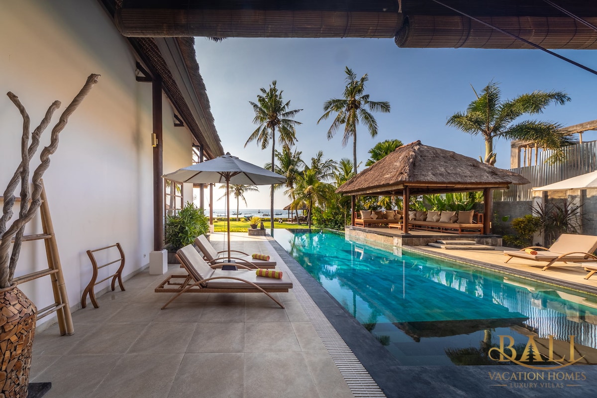 An inviting infinity pool reflects the clear blue sky, bordered by a tiled deck featuring sun loungers and umbrellas. Lush palm trees provide shade, and a traditional Balinese pavilion can be seen in the background, highlighting the serene outdoor setting of the villa.