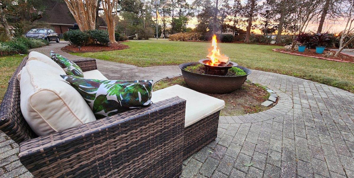 An outdoor seating area is featured with a comfortable couch adorned with tropical-patterned pillows. A circular fire pit is set in the center, surrounded by a landscaped area. The lush greenery and colorful plants enhance the natural beauty of the surroundings, with a soft glow from the sunset in the background.