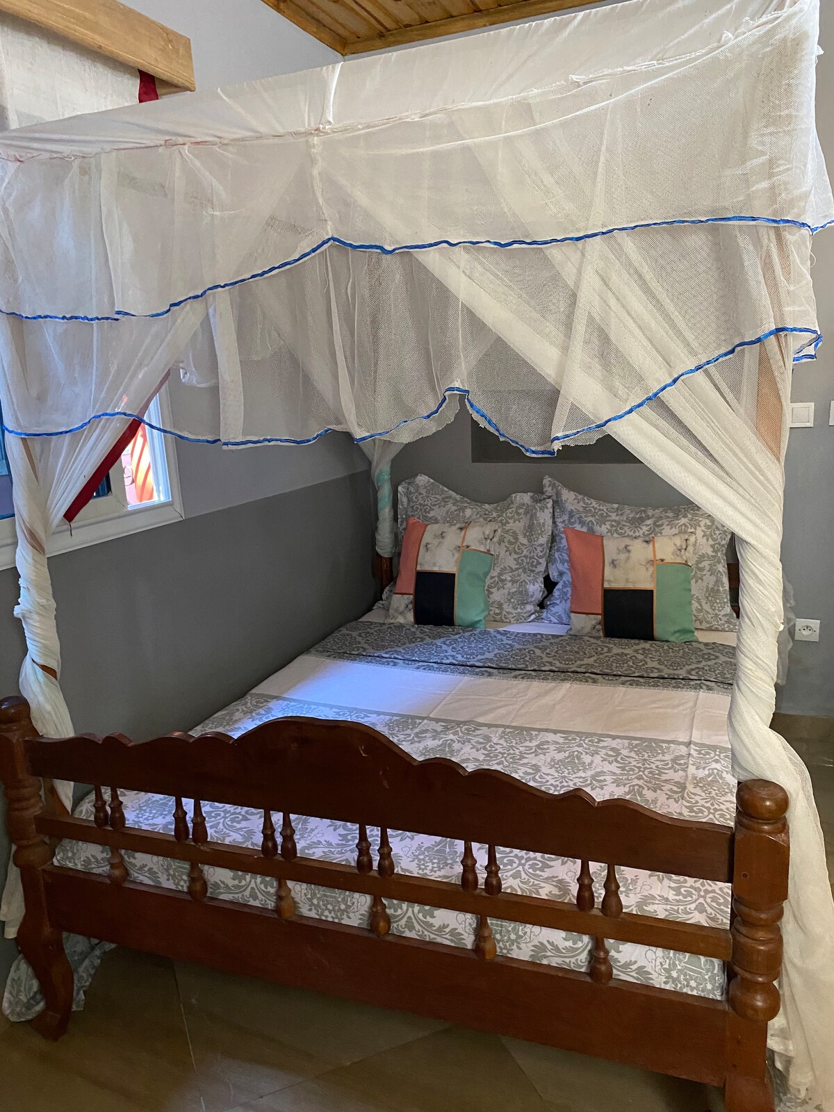 A cozy sleeping area features a wooden bed with a decorative canopy. The bed is dressed with patterned linens and accent pillows in soft colors. Natural light filters through a nearby window, which is framed by neutral-colored walls.
