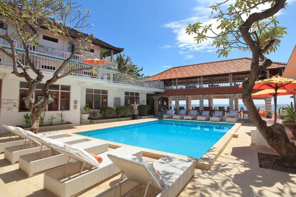A communal pool area is framed by lounge chairs and tropical trees, providing a bright and open atmosphere. The square-shaped building surrounds the pool, while a covered seating area overlooks the water, creating a relaxed setting for social gatherings.