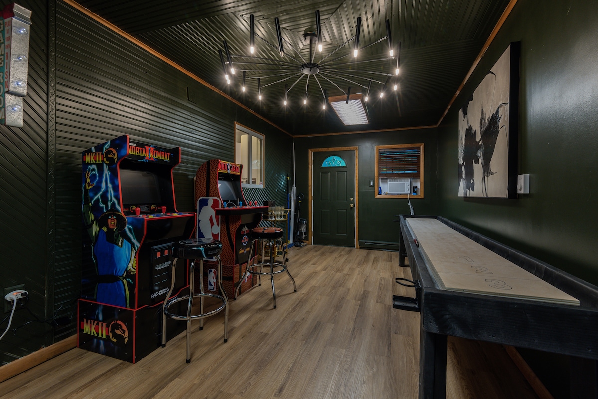 The game room features vintage arcade machines, a shuffleboard table, and bar-style seating. A modern chandelier provides overhead lighting. The dark green walls create a cozy atmosphere, while a door leads to the outside, and a large artwork decorates the side wall.