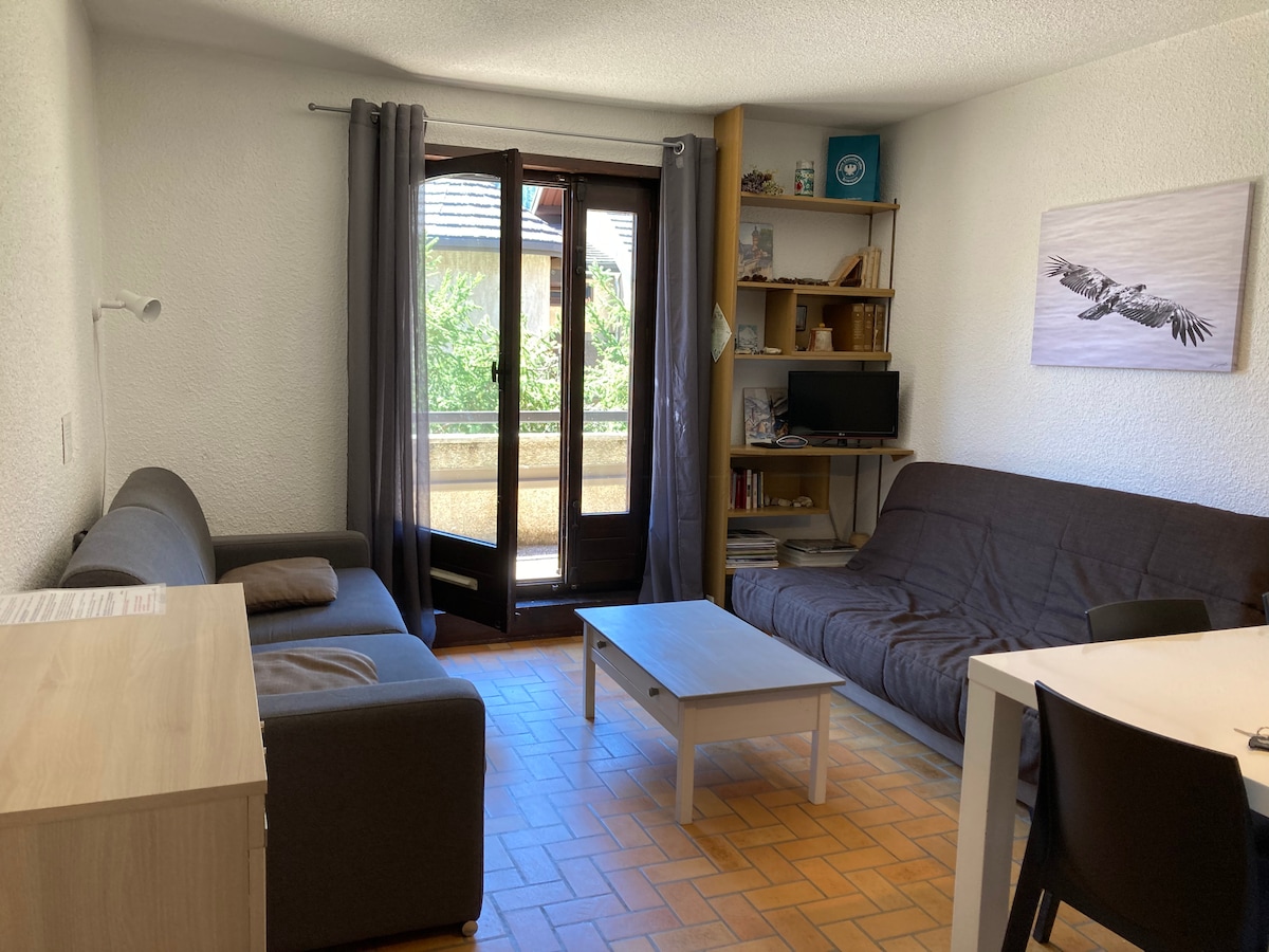 A living area is displayed, featuring a gray sofa and a brown futon. A light wood table sits at the center, surrounded by a black chair. Large doors open to a balcony, allowing natural light to illuminate the space.