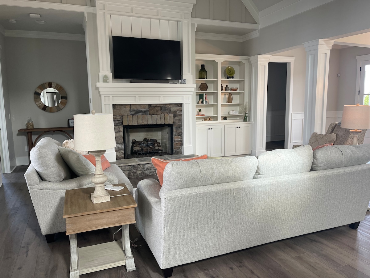 A cozy living area is presented, featuring a light grey sectional sofa with coral accent pillows. A wooden side table holds a lamp next to the seating. A stone fireplace serves as a focal point, with a mounted television above it. Decorative shelving units are visible in the background.