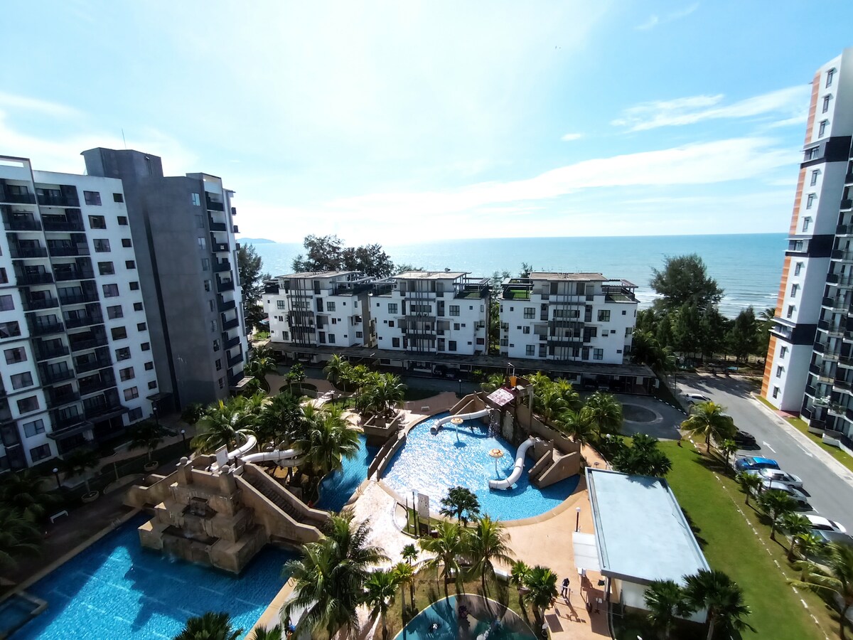 A sweeping view of the resort showcases the swimming pool and mini water theme park, surrounded by lush palm trees. The South East China Sea glimmers in the background, with spacious buildings lining the beachfront, creating a tranquil atmosphere.