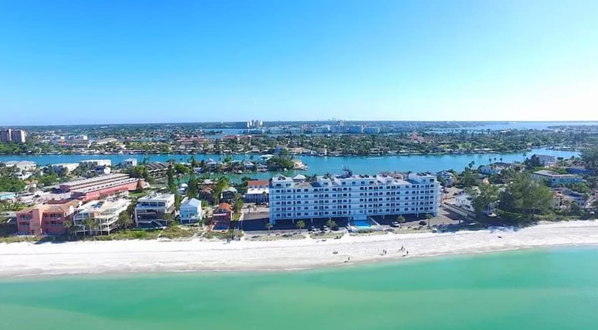An aerial view captures the spacious beachfront property bordered by crystal-clear waters and soft sand. The five-story building features a sunlit exterior and is surrounded by lush green landscapes, with nearby residential areas and waterways visible in the background.