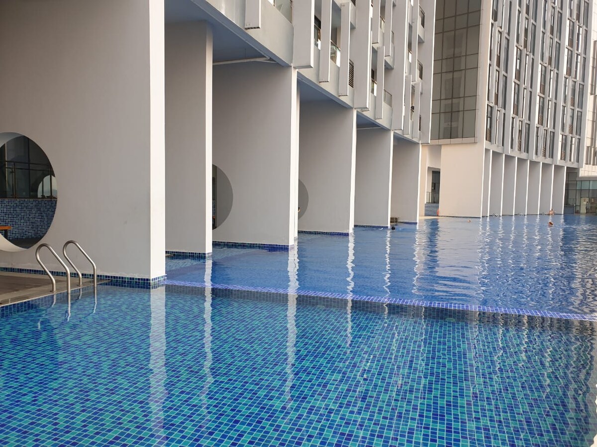 A spacious outdoor pool is seen with clear blue water, reflecting the surrounding modern architecture. White columns create a geometric pattern, and a set of stairs leads into the pool, offering easy access. The tranquil environment encourages relaxation.