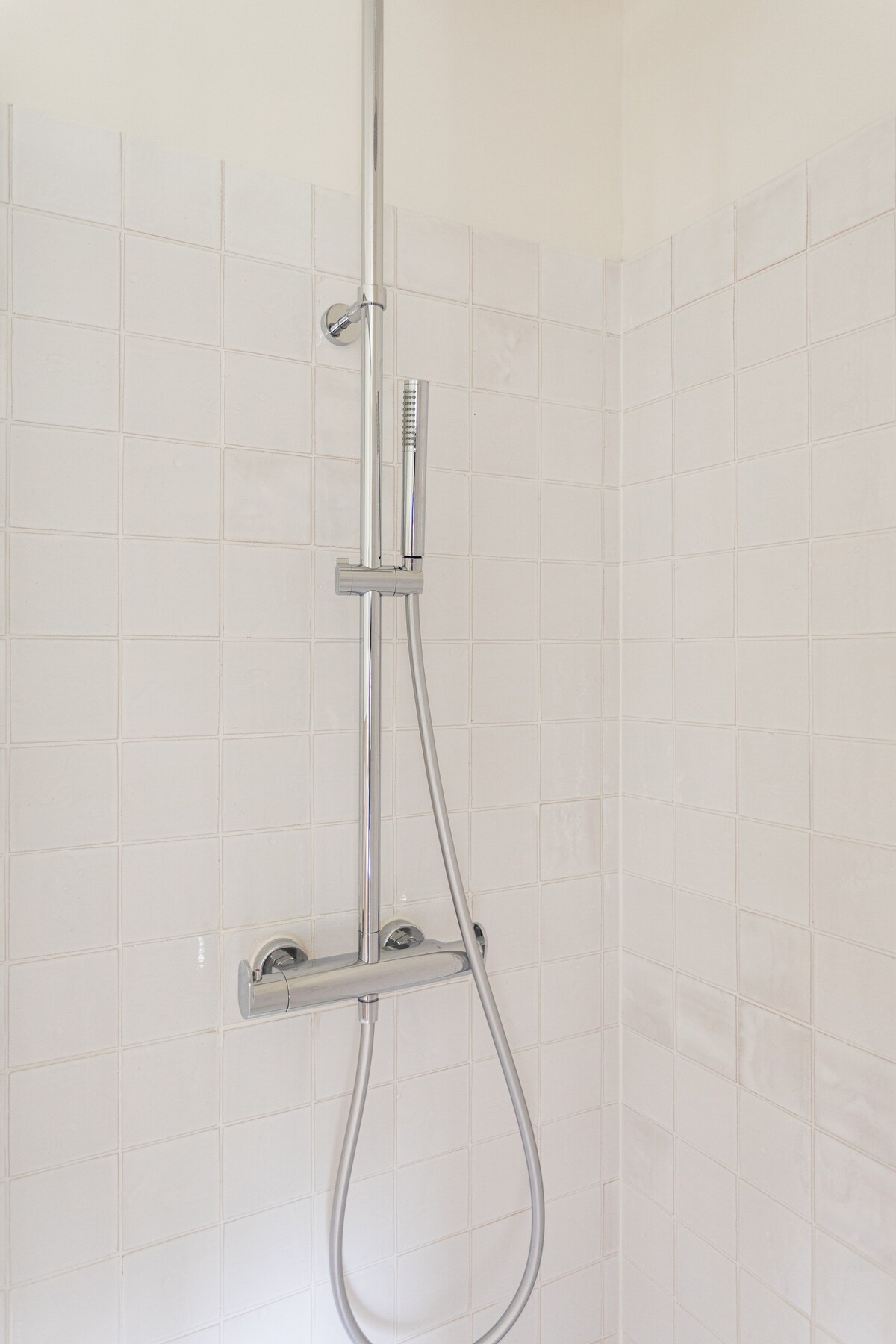 A modern shower area features sleek chrome fixtures against a backdrop of white tiled walls. The design includes a handheld showerhead and a minimalist shower control panel, providing a clean and simple aesthetic.