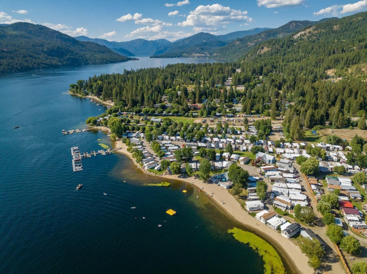 An aerial view captures Christina Lake and the surrounding area, showcasing a blend of lush greenery, sandy shores, and serene waters. The layout of the park model RVs is visible, positioned near the beach, with a backdrop of rolling hills and a clear blue sky.
