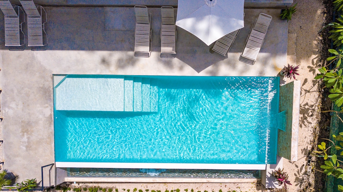 An aerial view showcases a serene infinity pool surrounded by a spacious patio. Several lounge chairs are positioned beneath a large umbrella. The clear blue water sparkles under sunlight, highlighting the pool's clean lines and inviting atmosphere.