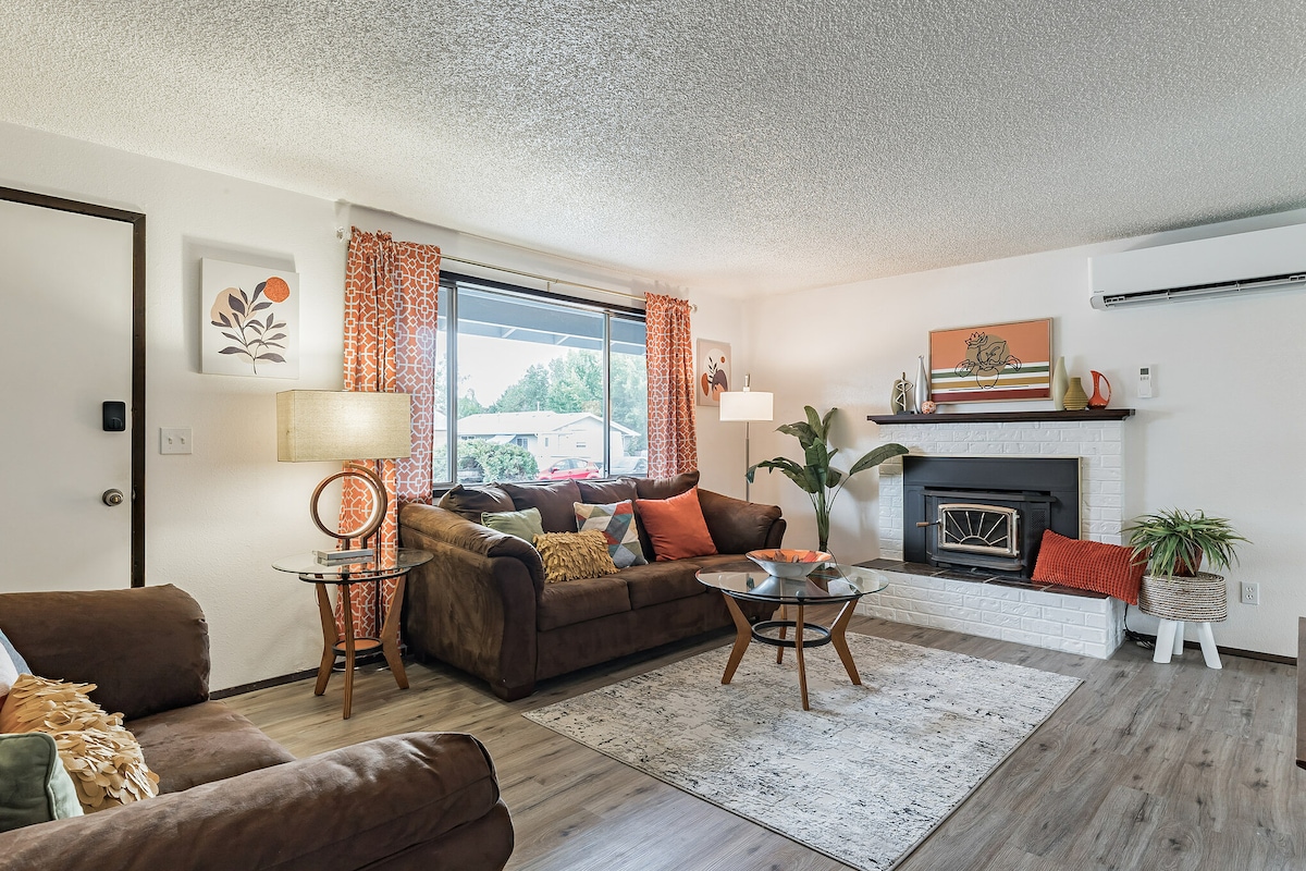 The living room features a cozy seating arrangement with a brown couch and armchair, complemented by colorful throw pillows. A glass coffee table stands on a light area rug, while a fireplace acts as a focal point. Large windows allow natural light to fill the space.