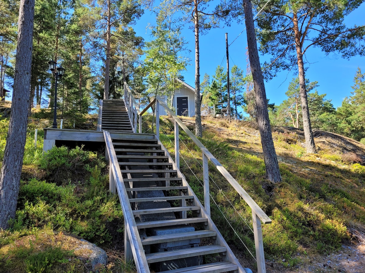 A wooden staircase ascends through a forested area, flanked by trees and greenery. At the top of the stairs, a cottage is visible, positioned among the natural landscape. Clear blue skies provide a bright backdrop, enhancing the tranquil setting.