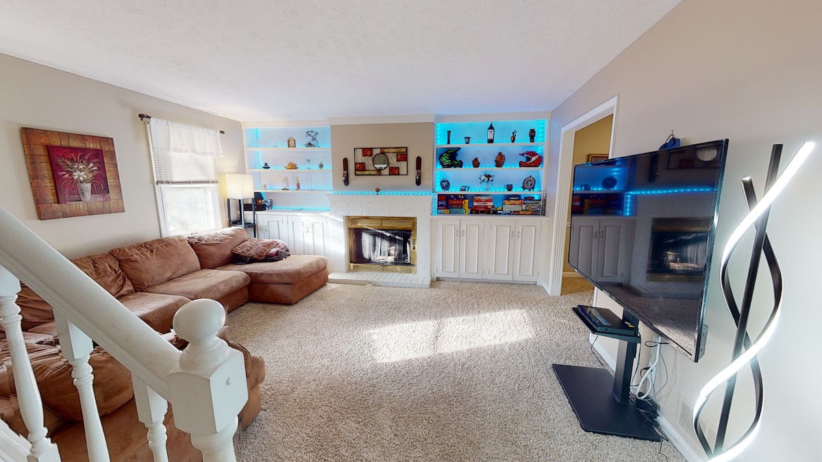 A cozy living room is featured, showcasing a large sectional couch and a flat-screen TV on a sleek stand. Built-in shelves with colorful items provide visual interest, while a fireplace offers warmth. Natural light illuminates the soft carpeted floor, enhancing the space's inviting feel.