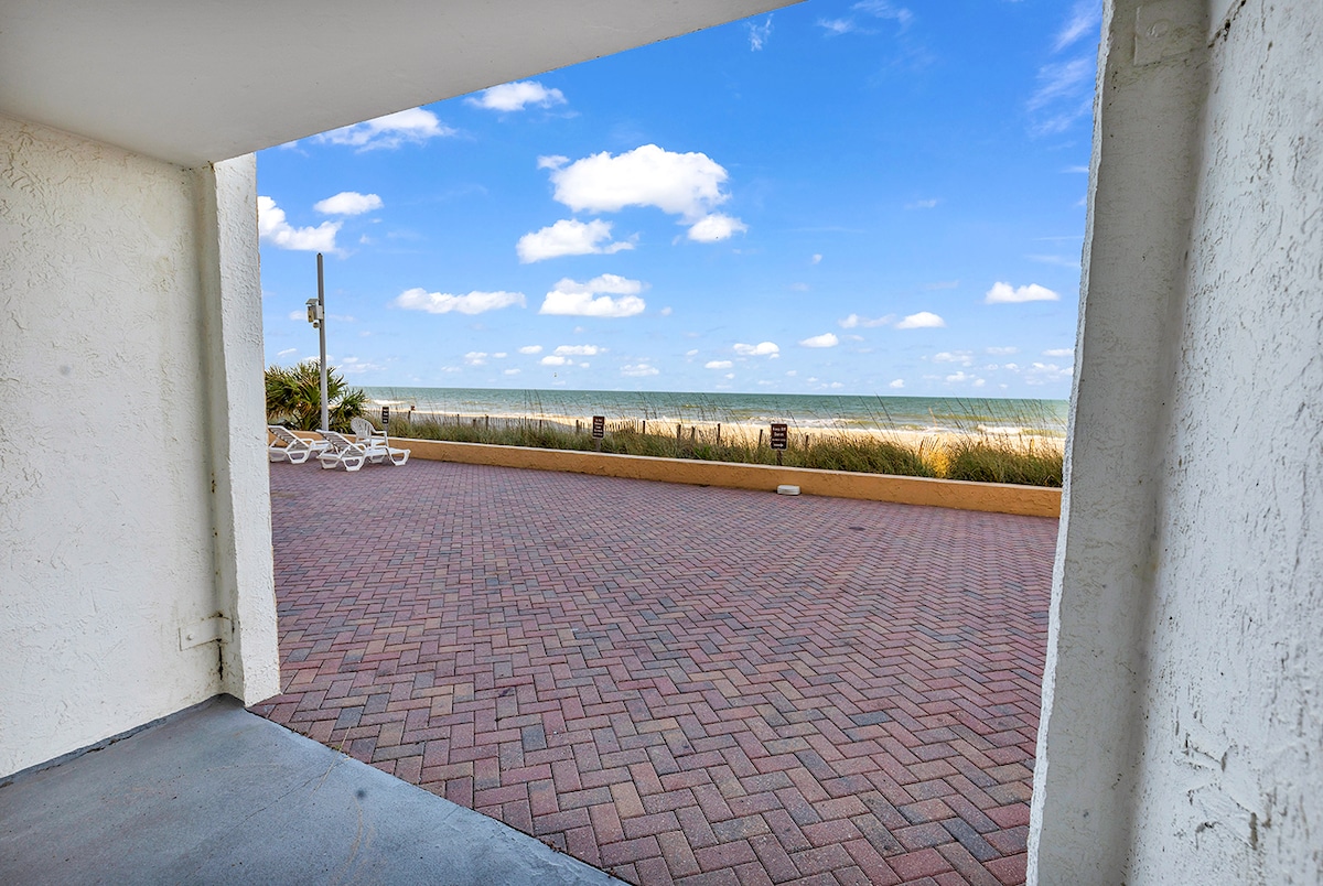 The view from the private patio showcases a wide stretch of sandy beach and the ocean under a bright sky. Soft clouds drift above, and beach chairs are visible in the distance, providing a glimpse of leisure near the water.