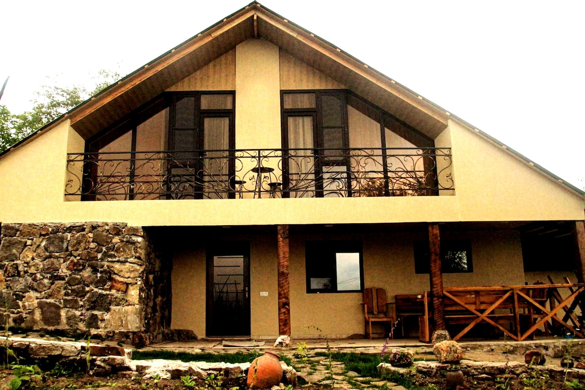 The two-story guest house features a welcoming façade with large windows and a balcony adorned with decorative ironwork. Natural stone elements are present at ground level, complemented by a spacious outdoor area with wooden seating, surrounded by plant life.
