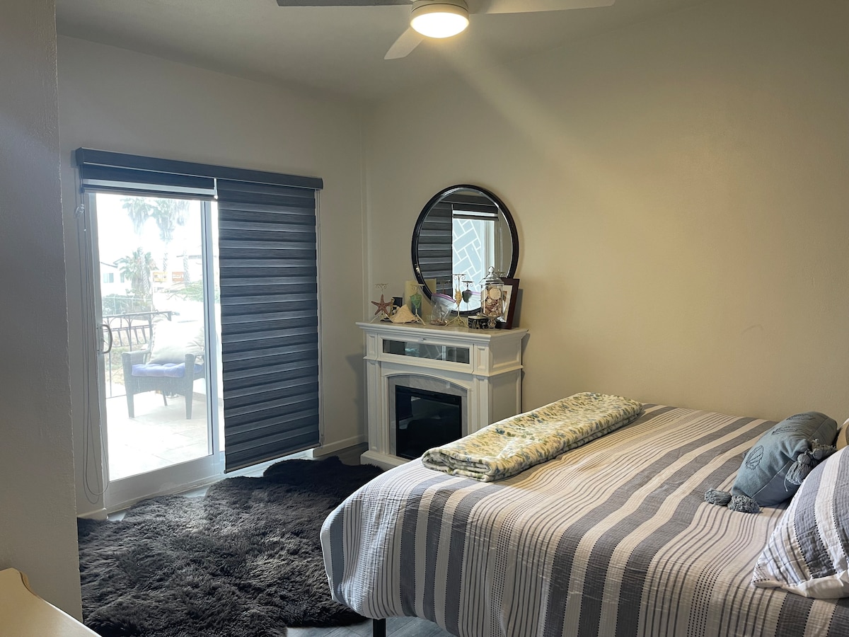 A bright sleeping area features a spacious bed covered with a striped blanket. A cozy area rug is positioned underneath. A circular mirror hangs above a decorative mantel. Large windows offer natural light, with sliding shades partially drawn, creating a serene atmosphere.