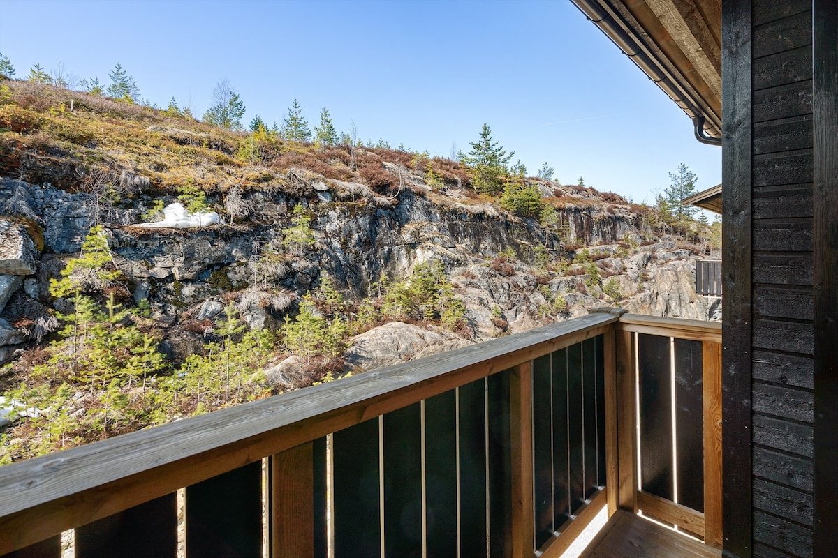 The balcony offers a view of rugged terrain, featuring rocky outcrops and sparse vegetation. Pine trees dot the landscape, creating a natural backdrop that emphasizes the tranquility of the surroundings.