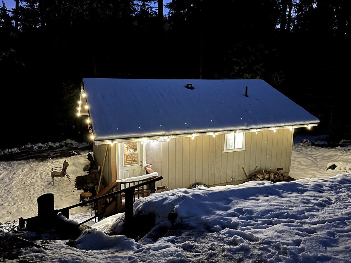 A cozy cabin is seen nestled in a snowy setting, illuminated by soft lights along the roofline. Steps lead from a wooden platform to the entrance, surrounded by a blanket of snow. The dimly lit forest provides a serene backdrop.