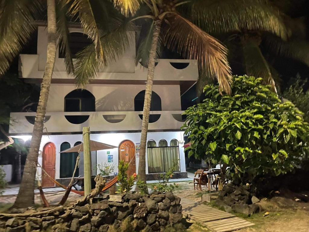 A multi-story building with a white exterior is surrounded by tropical palm trees and lush greenery. Softly lit by night, the structure features arched windows and wooden doors. A small patio area with dining furniture is visible in front, enhancing the relaxed atmosphere.