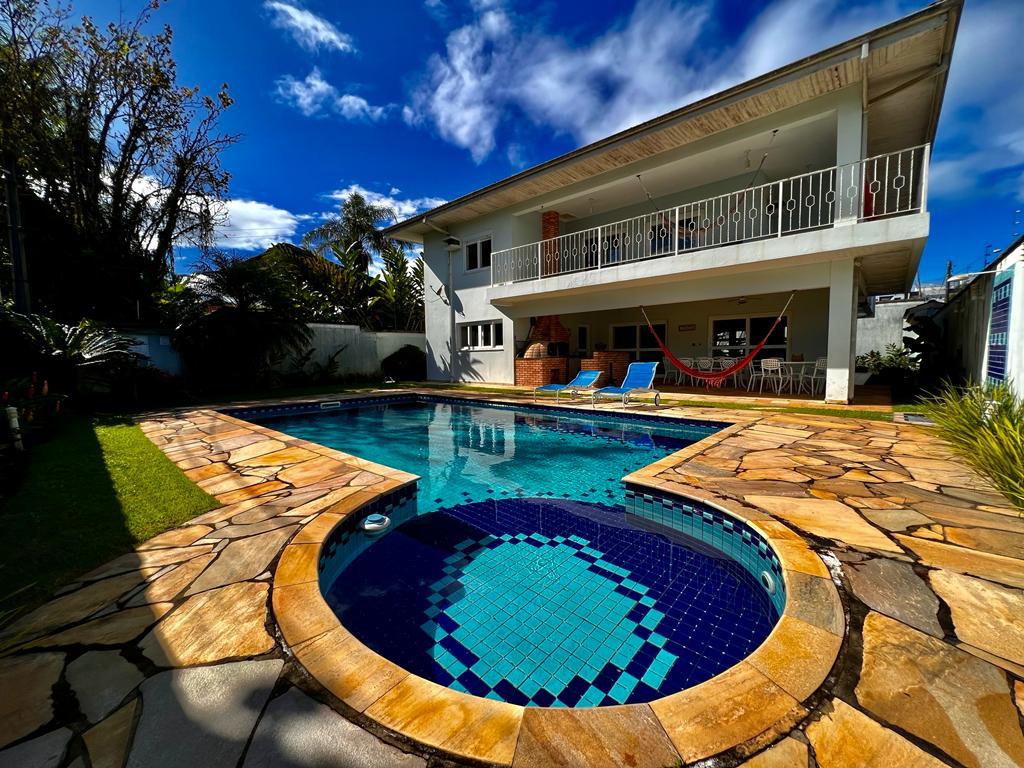 A spacious outdoor area features a rectangular swimming pool surrounded by natural stone decking. Lounge chairs are arranged beside the pool, while a hammock is suspended on a covered terrace above. Lush greenery and open sky provide a relaxing atmosphere.